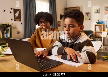 Black child and Black teenager studying on laptop at home, following online lesson and doing homework. Useful for e learning, homeschooling, digital education, medium shot Stock Photo