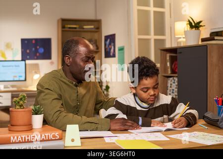 Black senior man helping Black boy studying and writing in workbook at home, supporting homework and learning. Useful for parenting, tutoring, education support campaigns Stock Photo
