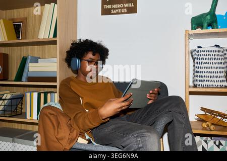 Black teenager wearing headphones using tablet while sitting in chair at home, engaging in online learning or digital entertainment. Ideal for education technology, screen time, youth media use Stock Photo