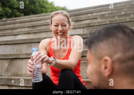 Smiling woman resting with a water bottle after an outdoor workout session. Sporty woman sitting on urban stairs and talking with her training partner Stock Photo