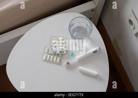 Pills are placed in their packaging on a white table along with a glass of water. A medical device is next to the pills, all in a room with a bed near Stock Photo