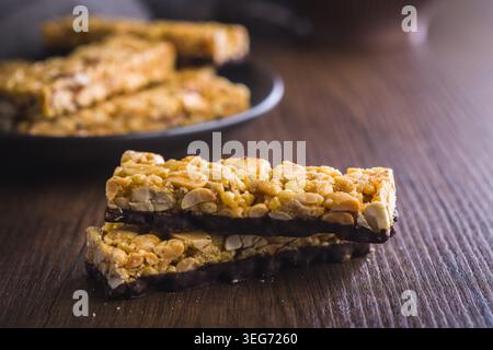 Stacked peanut granola bars with a dark chocolate base on a rustic wooden surface, close-up shot highlighting crunchy oats, peanuts and sweet caramel for a tasty snack or breakfast treat. Stock Photo