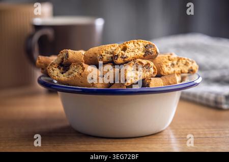 Homemade Italian biscotti cantuccini piled in an enamel bowl and scattered on a rustic wooden table, ideal for coffee or tea breaks. Warm, crunchy golden baked cookies that evoke cozy mornings and casual hospitality. Stock Photo