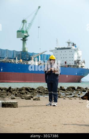 Female maritime engineer using digital tablet for logistics management and industrial port operations near cargo vessel Stock Photo