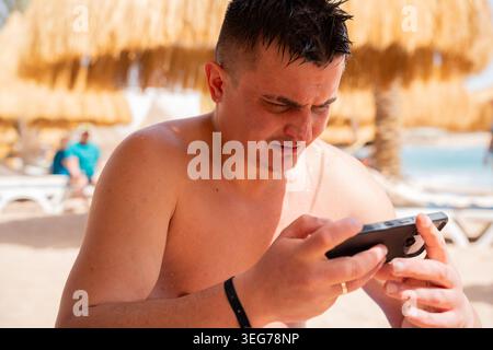 Caucasian man using smartphone at beach resort in Sharm El Sheikh Egypt near Red Sea umbrellas. Remote work and travel lifestyle. Stock Photo