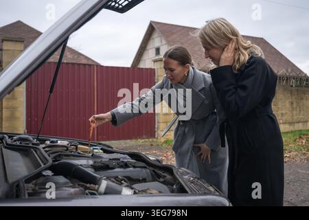 Two women standing next to a broken down car with its hood open, one checking the engine oil level with a dipstick, while the other looks concerned ab Stock Photo