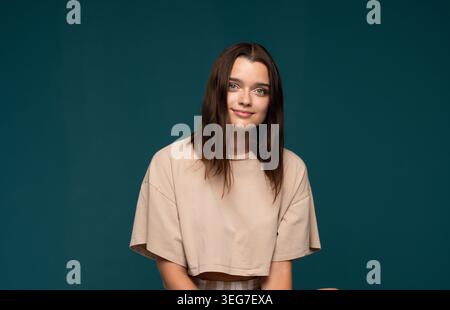 Young woman confidently smiling, posing against a clean blue-green background, expressing natural casual beauty and happiness Stock Photo