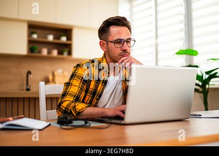 A freelancer studies something on his laptop, surrounded by a notebook and desk items in a modern home workspace. Stock Photo