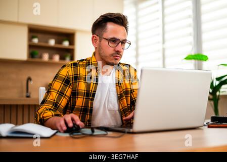 A freelancer studies something on his laptop, surrounded by a notebook and desk items in a modern home workspace. Stock Photo