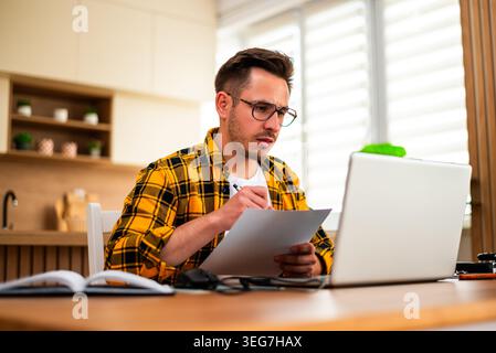 A young man sits at a wooden table, focuses on document while using laptop for freelance work. Stock Photo