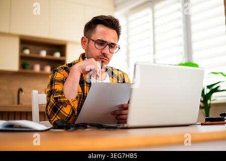 A young man in glasses sits at a wooden table, focuses on document while using laptop for freelance work. Stock Photo