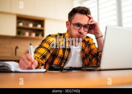 A frustrated remote worker struggles to concentrate, holding a pen while staring at his laptop in a home office environment. Stock Photo