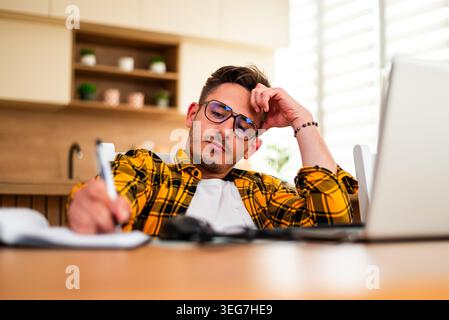 Man studies his laptop screen while writing in a notebook, suggesting pressure from work or study. Stock Photo