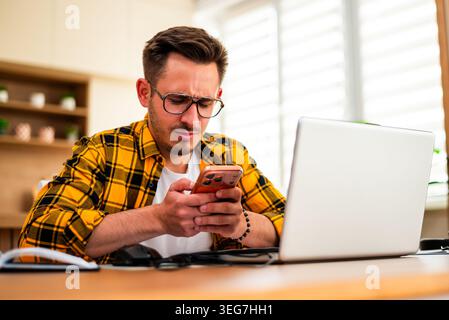 With a tense expression, a man scrolls through his phone at his desk, suggesting stress from work or unexpected news. Stock Photo