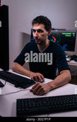 Vertical portrait of man focused on computer work late at night. Moody lighting from monitors highlights concentration. Concept of cybersecurity Stock Photo
