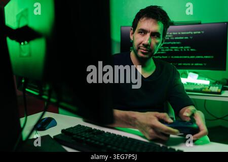 Male software developer playing video games after programming, sitting in dark room illuminated by green light from computer screens, mixing work and Stock Photo