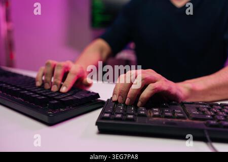 Unrecognizable programmer hands typing on two computer keyboards in dark interior of room with purple light, concept of code development, data entry Stock Photo