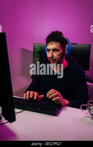 Vertical portrait of young man with headphones works on computer in dark room with pink neon light. Programmer coding or gamer playing video game Stock Photo