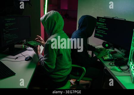 Two anonymous hackers in hoodies collaborating on cybercrime. Programmers work late at night in dark room with green light. Concept of cybersecurity Stock Photo