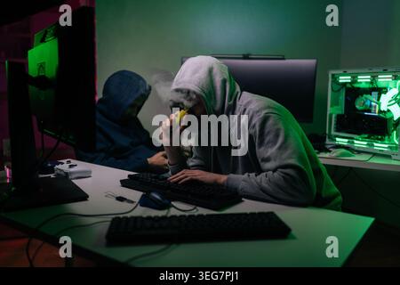 Two anonymous hackers in hoodies work on computers in dark room. One person vapes coding. Green and pink neon lights from gaming equipment create Stock Photo