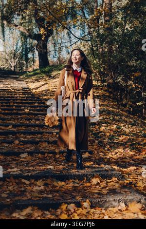 Woman walks down stone steps in autumn park holding leaves. Celebrating autumn seasonal changes, optimism and well-being, authentic happy expressions. Stock Photo