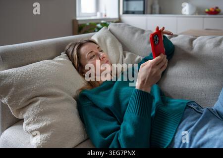 Calm midlife woman using phone home rest on sofa enjoys convenient online shopping, scrolling webs. Stock Photo