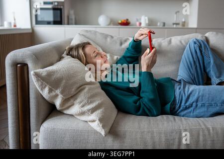 Calm midlife woman using phone enjoy online shopping, scrolling social media resting on home couch  Stock Photo