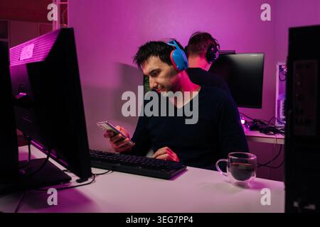 Young man with headphones looking at smartphone sitting at computer. Another person in background. Room lit with modern purple neon lights, creating Stock Photo