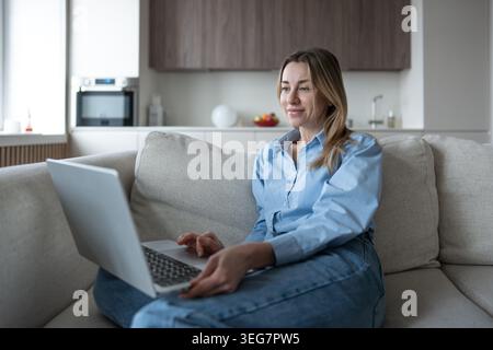 Contented middle aged woman using laptop relaxed sitting at home couch interested looks at screen. Stock Photo