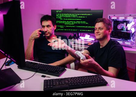 Two young men at desk with computers. One programming code on screen other plays video game with controller. Scene illuminated by purple neon lights Stock Photo