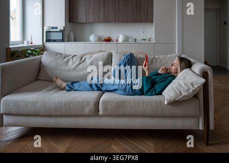 Contented middle aged woman using phone lounging on sofa online shopping with interest from home. Stock Photo