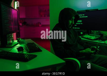 Mysterious programmer in black hoodie works late, surrounded by computer monitors showing code, room lit by dramatic green and pink neon light Stock Photo