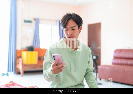 A lifestyle portrait of a handsome young Asian man from the Isan region of Thailand sitting indoors and looking at his mobile phone. He focuses on the Stock Photo