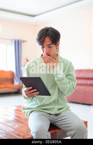 A vertical lifestyle portrait of a shocked young Asian man from the Isan region of Thailand sitting indoors on a wooden bench while using a tablet com Stock Photo