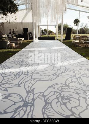 Elegant floral pattern adorns a long white carpet. Chandelier-like drapes hang from a modern canopy. Outdoor wedding venue under open sky. Natural lig Stock Photo