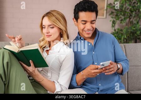 Couple relaxing on sofa, woman reading book man using smartphone Stock Photo