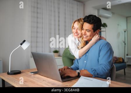 Happy couple working from home using laptop embracing Stock Photo