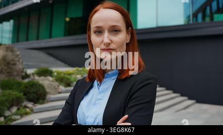 Portrait Caucasian serious confident businesswoman posing hands crossed outdoors moving shot in city girl looking at camera business female woman real Stock Photo