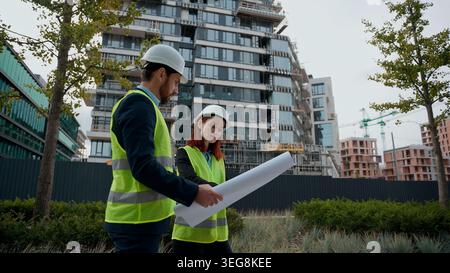 Two architects build workers Caucasian woman man engineers discuss plan building female male colleagues partners in safety hat reflective vest talking Stock Photo