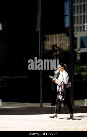 A woman walks on the sidewalk near a glass building. She looks at her phone while wearing a light shirt and dark pants. The sun shines brightly, creat Stock Photo