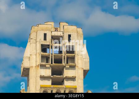 Tall abandoned concrete tower with unfinished brutalist architecture standing against a vivid blue sky with soft clouds in an urban industrial environ Stock Photo