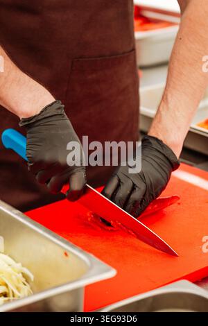 Close-up of chefs hands in black gloves skillfully slicing with a large chef knife on a bright red cutting board. Stock Photo