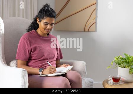 Asian woman sitting and writing in spiral-bound notebook on lap in home lounge, wearing maroon tee Stock Photo
