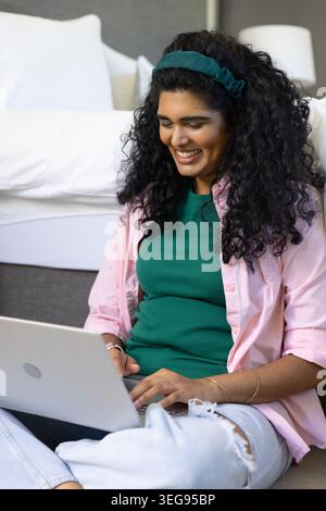 Indian woman sitting on bedroom floor using laptop, wearing green top ripped jeans headband Stock Photo