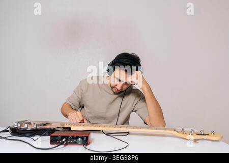 Thoughtful man wearing headphones and concentrating intensely while resting head on hand, sitting at desk with bass guitar and red audio interface Stock Photo