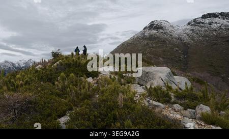 Two hikers standing on a rugged mountain ridge looking at the view with snow-covered peaks under a dramatic cloudy sky in the southern Andes of Chile. Stock Photo