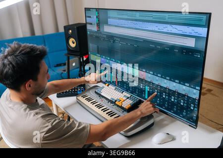 Rear view of sound engineer male creating new music in home studio, arranging tracks and mixing audio levels with midi keyboard and software on a Stock Photo