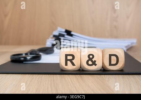 Close-up photo of wooden cubes spelling R and D placed on a black folder on a wooden office desk. Blurred background includes stacked documents and a Stock Photo