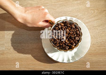 Hand Holding Cup of Roasted Coffee Beans. Hand holding an elegant cup filled with roasted coffee beans on a saucer, lit by warm sunlight. Stock Photo