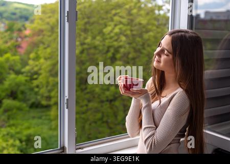 Woman Drinking Tea by Open Window. Young woman holding a red cup by an open window, enjoying a peaceful moment with green nature outside. Stock Photo